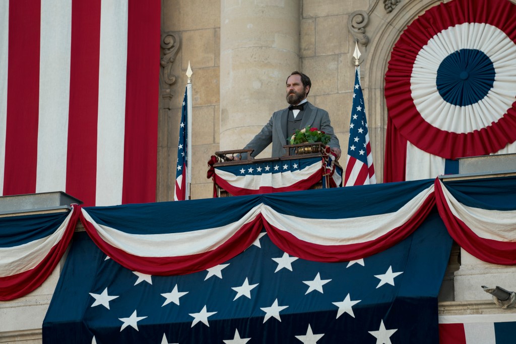"A man in historical attire stands at a decorated podium on a balcony, surrounded by American flags and patriotic bunting, evoking a formal and patriotic atmosphere."