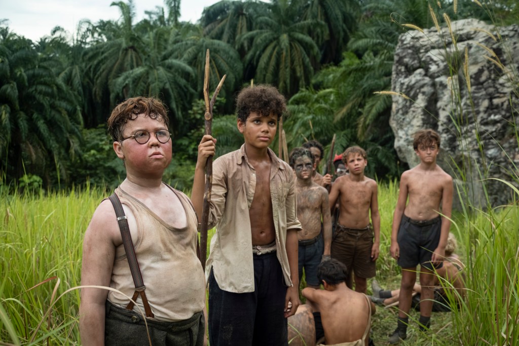 "A group of dirty, shirtless boys stand in tall grass with jungle foliage and a rock behind them. Some hold makeshift spears, appearing serious and alert, as if stranded or surviving in the wild."